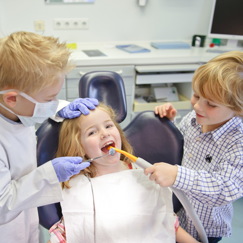 Kinder spielen Zahnarzt: ein M&auml;dchen sitzt lachend im Behandlungsstuhl, zwei Jungen halten Mundspiegel und Absauger